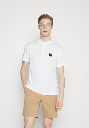 Polo bianco a maniche corte, con colletto, tre bottoni e un patch logo nero sul petto. Indossato con pantaloni corti beige.