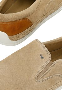 Beige suede slip-on shoes with stitched brown leather accents, white soles, and a small metallic logo on the upper side.