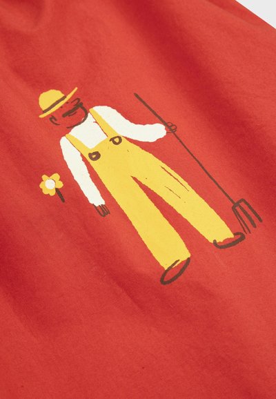 Homme en salopette jaune et chapeau tenant une fourche à côté d'une petite fleur jaune sur un fond en tissu rouge.