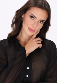 Musta läpikuultava pusero, jossa on pitkät puffihihat, rypytetyt kalvosimet ja valkoiset napit. Pyöreä pääntie ja sileä pinta.