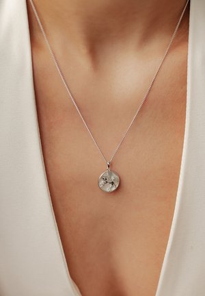 Silberne Anhänger-Halskette mit einem kreisförmigen Design und durchbrochenen Blumenmustern, aufgehängt an einer feinen Kette, vor einem hellen Stoffhintergrund.