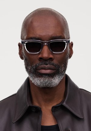 Sonnenbrille mit einem klaren Rahmen und dunklen Gläsern, im rechteckigen Design. Der Träger hat einen kurzen grauen Bart und eine kahle Kopf.