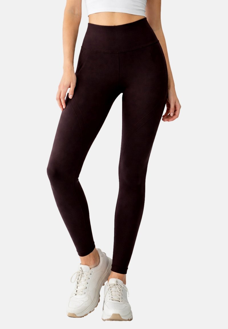 Femme portant des leggings noirs taille haute et des baskets blanches épaisses, debout les jambes croisées devant un fond uni.