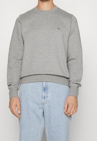 Szara bluza z długimi rękawami, ściągaczami na mankietach i dole, z małym logo na piersi. Połączona z jasnoniebieskimi dżinsami.