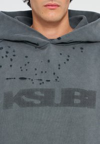 Sivá mikina s kapucňou s opotrebovaným dizajnom a malými dierkami, na prednej strane s čiernym potlačeným logom "KSUBI". Mäkká textúra.