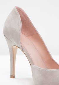 Chaussure à talon haut en daim gris clair avec un bout pointu, un talon fin et une doublure intérieure rose pâle sur un fond blanc.