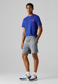 Μπλε κοντομάνικο μπλουζάκι με το λογότυπο "Red Bull", συνδυασμένο με γκρι αθλητικές βερμούδες με ελαστική μέση και πλαϊνές τσέπες. Άσπρες κάλτσες αστραγάλου, πολύχρωμα αθλητικά παπούτσια.