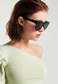 Mustat aurinkolasit, joissa on geometrinen kehys, matta pinta ja tumma linssi. Yhdistetty vaaleanvihreään yhden olkapään yläosaan, sileä kangaspinta.