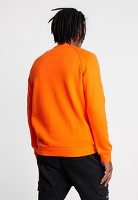 Oranžová mikina s kulatým výstřihem, dlouhými rukávy a žebrovanými lemy na rukávech. Látka vypadá měkká s hladkou strukturou.