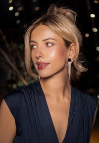 Robe bleu marine avec texture plissée, boucles d'oreilles à franges argentées et décolleté vertical. Un éclairage doux et chaud met en valeur les traits.