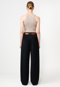 Naine seisab seljaga kaamera poole, seljas beež ilma varrukateta topp, mustad laiad püksid ja pruun vöö, juuksed madalas pundis.