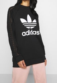 Svart sweatshirt i bomull med en stor vit Adidas-logotyp och randiga snördetaljer på axlarna. Långa ärmar och ribbad mudd.