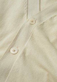 Tissu tricoté de couleur crème avec deux boutons assortis cousus sur une patte verticale chevauchante, texture fine côtelée et détails de couture subtils.