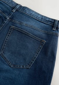 Sötétkék farmernadrág, egyetlen hátsó zsebbel, amely téglalap alakú, kontrasztos varrással és sima, textúrált felülettel.