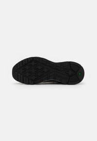 Semelle de chaussure en caoutchouc noir avec un motif de bande de roulement en forme de croix et un petit détail hexagonal vert près du talon sur fond blanc.