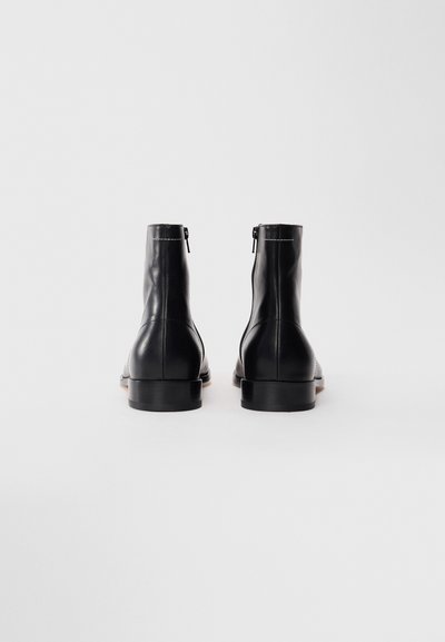 Ein Paar schwarze Leder-Ankle-Boots von hinten gezeigt, mit Reißverschluss und niedrigen Blockabsätzen auf einem weißen Hintergrund.