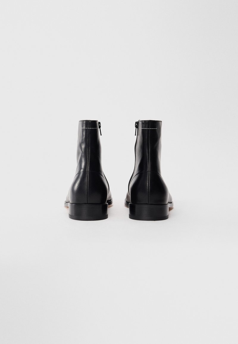 Une paire de bottes en cuir noir à lacets montrée de derrière, avec des fermetures éclair et des talons bas empilés sur fond blanc.