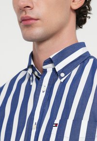 Μπλε και άσπρη ριγέ πουκάμισο με κουμπωτή γιακά, μπροστινή τσέπη με λογότυπο, φτιαγμένο από ελαφρύ ύφασμα.