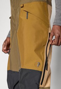 Közelkép egy ember kezéről, ahogy kinyitja a oldalsó cipzárt színes blokkos szabadtéri nadrágon, amely barna, mustár és fekete panelokból áll.