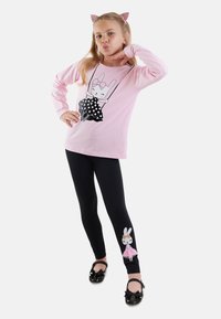 Rosa Langarmshirt mit einem Hasenmotiv in einem gepunkteten Kleid, kombiniert mit schwarzen Leggings, die am Knöchel ein Hasen-Design haben. Schwarze Schuhe.