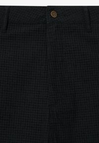 Mustat housut, joissa on hienovarainen ruutukuosi, valmistettu teksturoidusta kankaasta. Varustettu metallinapilla ja ommelluilla yksityiskohdilla saumoissa.