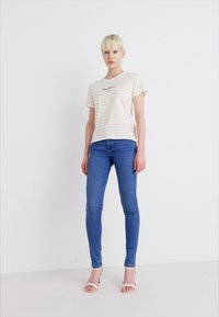 Camiseta de rayas en color beige claro con mangas cortas, combinada con unos vaqueros ajustados azules. La parte superior presenta un pequeño logo impreso en negro.