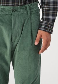 Groene corduroy broek met verticale ribbels, enkele knoopsluiting en elastische tailleband, gecombineerd met een geruit overhemd met lange mouwen.