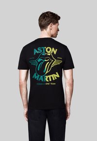 Czarny bawełniany T-shirt z kolorowym logo języka z skrzydłami, tekstem "ASTON MARTIN" i "FORMULA ONE TEAM" w żółtym i turkusowym kolorze.
