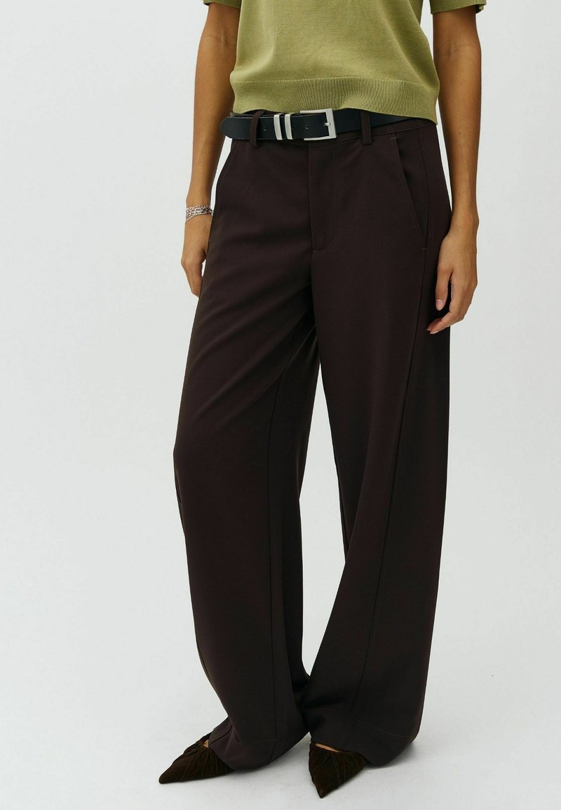 Personne portant un pantalon large foncé avec ceinture noire, un haut à manches courtes vert olive et des chaussures sombres pointues, debout les mains détendues le long du corps.