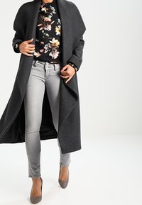 Lungo cappotto grigio in lana aperto su una blusa nera a fiori e jeans grigi a taglio aderente, completato da scarpe con tacco grigie.