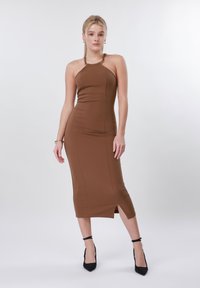 Γυναίκα με ξανθά μαλλιά, που φοράει ένα αμάνικο καφέ μίντι φόρεμα με πλαϊνή σχισμή και μαύρα τακούνια με λουράκι στον αστράγαλο, στέκεται μπροστά σε λευκό φόντο.