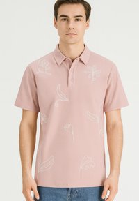 Lichtroze polo shirt met korte mouwen, ribbeltextuur en witte bloemenborduring. Beschikt over een kraag en een drieknoopsluiting.