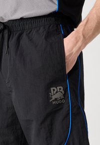 Pantalones cortos deportivos negros, fabricados con material ligero, con piping azul de acentuación y un logo gris en el muslo izquierdo.