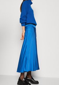 Sininen ribattuja korkeakaulusneule yhdistettynä pitkään, satiiniseen siniseen hameeseen. Hameessa on sileä pinta ja laskostettu helma. Mustat liukupohjaiset kengät.