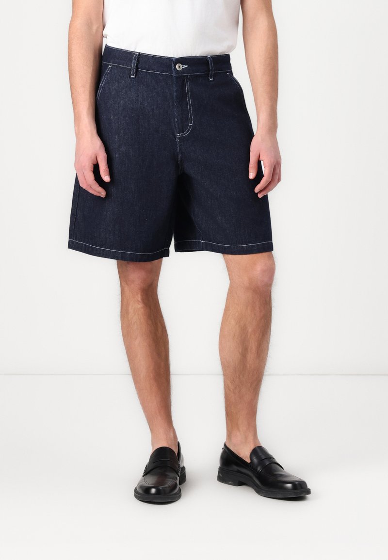 Mørke denimshorts med lige snit, der har hvidt stikning og en traditionel knaplukning. Bæres med sorte loafers.