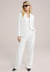 Completo bianco su misura con giacca aderente e pantaloni a gamba larga. Il modello indossa una maglietta bianca, occhiali da sole neri e scarpe nere a punta.