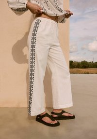 Pantaloni bianchi in lino a gamba larga con ricami neri lungo il lato, abbinati a sandali verde scuro con un design minimalista e cinturini regolabili.