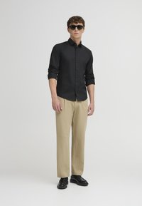 Schwarze Bluse mit Knöpfen, beiger Hose und schwarzen Schuhen. Das Model trägt Sonnenbrillen. Schlankes Design mit figurbetonter Silhouette und hochgekrempelten Ärmeln.