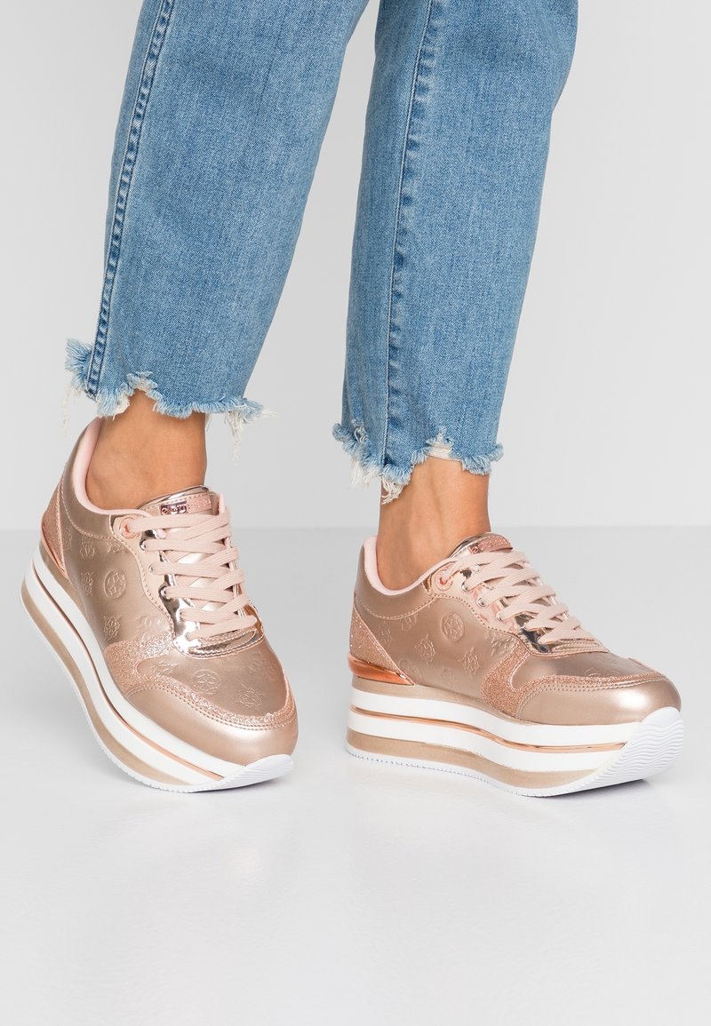 Baskets rose gold avec une finition métallique lisse, design à lacets et plateforme blanche épaisse. Présente des détails de logo subtils et des accents contrastants.