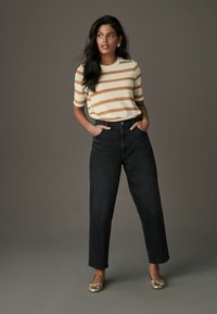 Gestreifter beiger und brauner Stricktop, hoch taillierte schwarze Jeans und metallische Ballerinas; Model steht mit Händen in den Taschen vor einem neutralen Hintergrund.