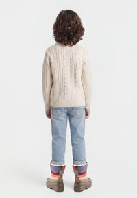 Enfant aux cheveux bouclés portant un pull crème à torsades, un jean bleu clair retroussé et des bottes d'hiver multicolores, debout dos tourné.