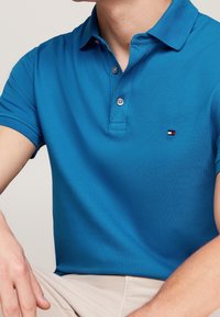 Tyrkysová polo košile vyrobená z texturované tkaniny, s předním zapínáním na tři knoflíky a malým logem poblíž hrudi.