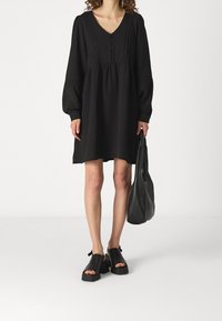 Robe noire à manches longues avec décolleté en V, détails de boutons et taille froncée. Associée à des sandales à talons bloc noires et à un sac à bandoulière.