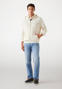 Beige Rippenjacke mit Reißverschluss und Kapuze, getragen über einem kragenbezogenen Hemd, kombiniert mit hellblauen Jeans und dunklen Schuhen mit braunen Schnürsenkeln.
