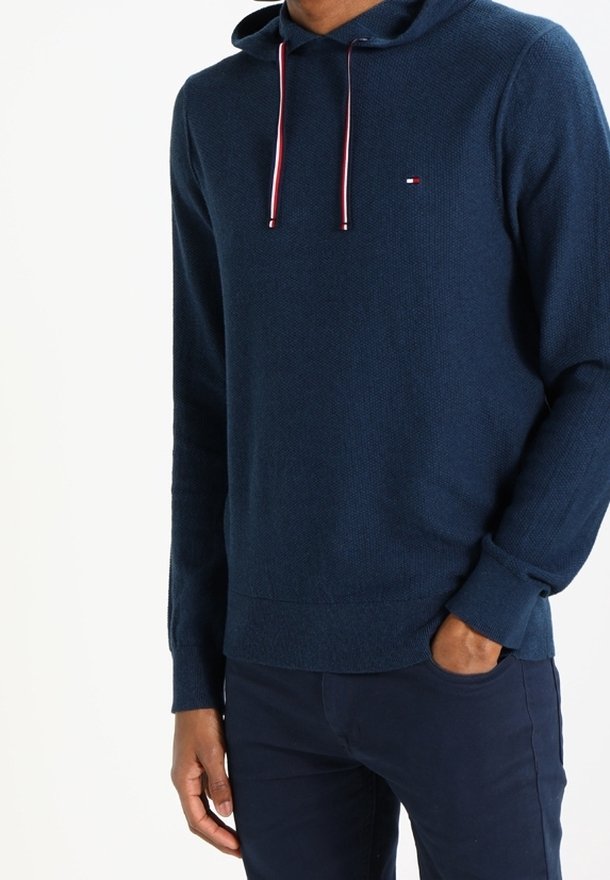 Hoodie lavorato a maglia blu navy con tessuto texturizzato, cordino rosso a contrasto e piccolo logo sul petto. Maniche lunghe e polsini a coste.