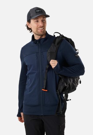 Uomo che indossa una giacca blu navy e un cappello nero, tiene la tracolla di uno zaino nero sulla spalla, guarda di lato e sorride.