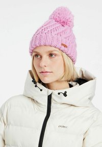 Roze gebreide beanie met een grote pompon bovenop en een textuurpatroon, gecombineerd met een witte gewatteerde jas met capuchon.