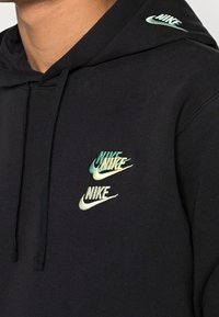 Svart hoodie med tre broderade Nike-logotyper i gradientnyanser av grönt och blått. Tillverkad av mjukt, texturerat tyg.