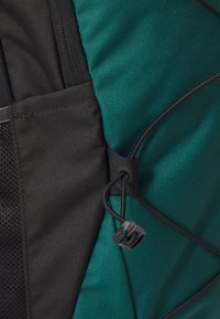 Material de mochila verde y negra con un acabado texturizado. Cuenta con un cierre de cordón y detalles de bolsillo de malla, mostrando correas ajustables y acentos.