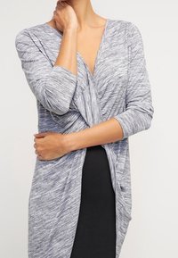 Šedý top s dlouhým rukávem a zkrouceným předním designem. Vyrobený z měkkého, strukturovaného materiálu, má hluboký výstřih do V a splývavou siluetu.